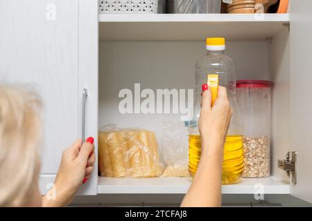 Die Hausfrau nimmt eine Flasche Sonnenblumenöl aus dem Küchenschrank. Frau, die eine Flasche Sonnenblumenöl hält Stockfoto