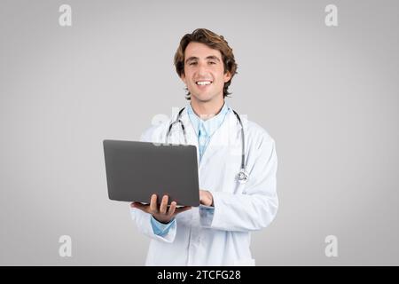 Medizinische Online-Forschung. Fröhlicher männlicher Arzt hält Laptop und lächelt in die Kamera, steht über grauem Hintergrund Stockfoto