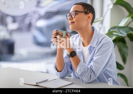 Entspannte Dame in Gläsern, die warmes Getränk genießen, mit einem ruhigen Lächeln und Notizbuch auf dem Tisch Stockfoto
