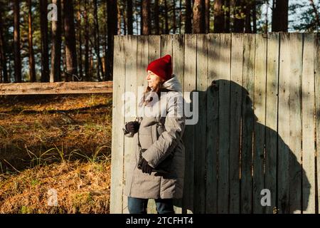 Eine Frau mittleren Alters strahlt herbstliche Eleganz aus, während sie an einem sonnigen Tag vor einem alten, schäbigen Zaun posiert und ihre stilvolle Herbstkleidung in einem Charme präsentiert Stockfoto