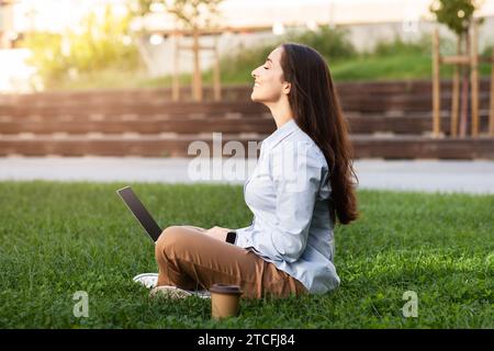Positive, ruhige, professionelle junge europäische Frau sitzt mit Laptop im grasbewachsenen Außenbereich Stockfoto