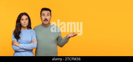 Schockierte kaukasische Männergesten mit offener Hand zur Erklärung neben ernster Frau mit gekreuzten Armen Stockfoto