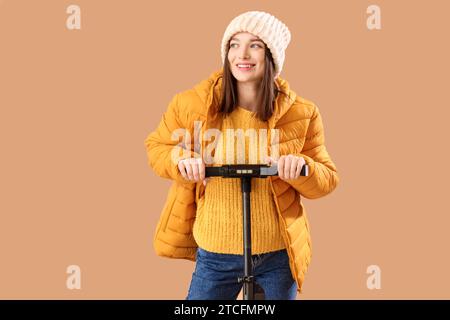 Junge Frau in Winterkleidung mit Elektroroller auf beigefarbenem Hintergrund Stockfoto