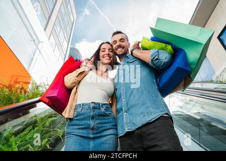 Ein junges echtes Paar, das auf der Rolltreppe des Einkaufszentrums mit Einkaufstaschen hinuntergeht. Verheiratete Kunden lächeln nach dem Kauf eines Bekleidungsverkaufs oder Rabatts auf die Stufen des Store Centers. Hochwertige Fotos Stockfoto