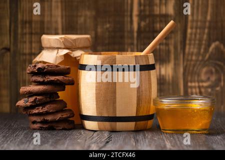 Honig in Gläsern und in einer Schüssel, Dipper und hausgemachte Kekse mit Schokoladenchips auf hölzernem Hintergrund. Zusammensetzung von handwerklichen Schokoladenkeksen und Honig Stockfoto