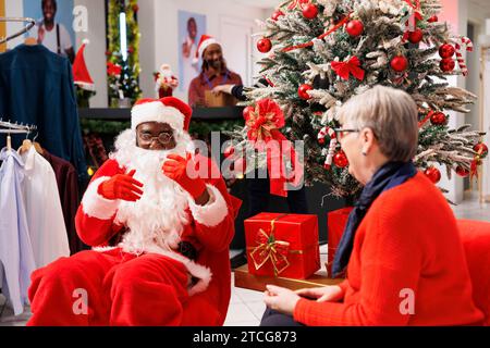 Der Mitarbeiter des weihnachtsmanns schenkt einer älteren Frau im Laden Geschenke und bietet eine festliche Kiste neben dem Weihnachtsbaum im Einkaufszentrum an. Ein leitender Mitarbeiter fühlt sich fröhlich, nachdem er Waren kostenlos erhalten hat, saisonale Verkäufe. Stockfoto