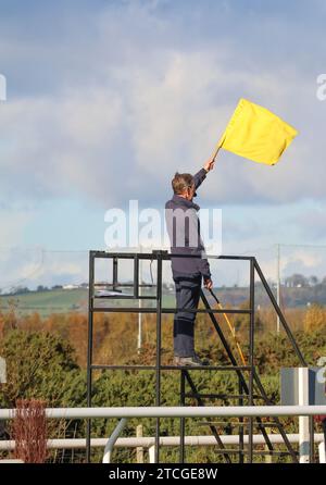 Start- und Startflagge UK National Hunt Race. Mann mit gelber Flagge startet Pferderennen unter Bestellung auf der Down Royal Racecourse, Lisburn, Nordirland. Stockfoto