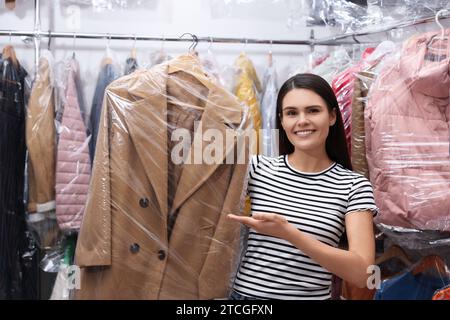 Reinigungsservice. Glückliche Frau, die einen Kleiderbügel mit Mantel in einer Plastiktüte in der Nähe von Regalen mit Kleidung im Haus hält Stockfoto