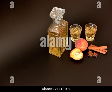 Pfirsichlikör mit Gewürzen auf schwarzem Hintergrund, eine Kristallkaraffe und zwei Gläser starken selbstgemachten Alkohol, Anis und Zimt daneben. Close-u Stockfoto