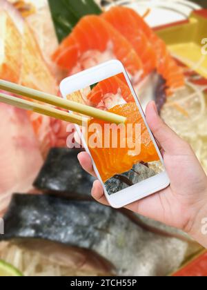 Weibliche Hand fotografiert Sashimi Sushi Set auf Sushi Bootsplatte mit Handy und Essstäbchen halten Lachs Sashimi im Handy Stockfoto