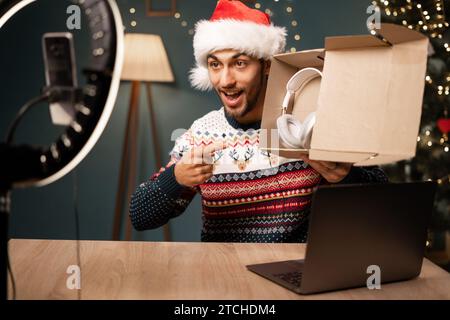 Arabischer männlicher Blogger, der ein Paket mit kabellosen Kopfhörern auf der Smartphone-Kamera auspackt, junger Influencer in Santa hat ein Produkt auspackt und ein Video filmt Stockfoto