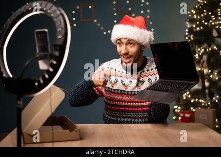 Junger arabischer Blogger, der weihnachtspaket mit einem neuen Laptop vor der Kamera entpackt und den Computer aus dem Karton nimmt, während er Videoinhalte für soziale Medien aufzeichnet Stockfoto