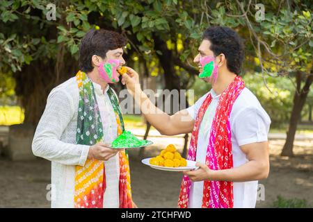 Zwei junge indische Freunde, die weiße Kurta tragen und gemeinsam holi feiern, während sie im Park oder Garten köstliche Laddu süße Süßigkeiten essen. Stockfoto