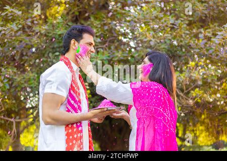 Glückliches, junges indisches Paar, das weiße Kurta spielt und Farbe ins Gesicht bringt. Feiern Sie gemeinsam holi im Freien im Park oder Garten. Stockfoto