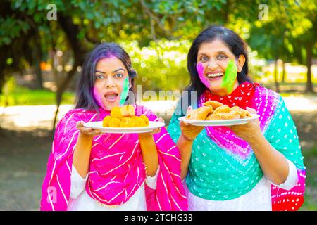 Zwei glückliche Indianerinnen tragen eine weiße Kurta mit einem Farbpuder oder gulal und Laddu, die das holi-Festival im Park oder Garten feiern. Stockfoto