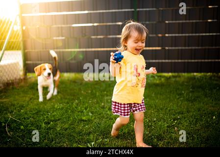 Baby-Mädchen, das mit Beagle-Hund im Garten läuft, am Sommertag. Haustier mit Kinderkonzept Stockfoto