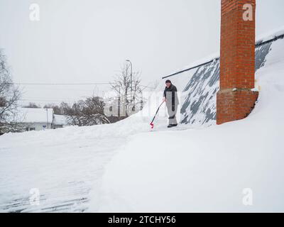 Ein älterer grauhaariger Dachreiniger entfernt an einem Wintertag Schnee vom Dach mit einer Schaufel. Schnee vom Dach eines Landhauses entfernen. Stockfoto