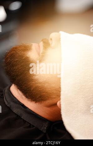Ein Kunde liegt auf einem Stuhl in einem Friseurladen mit seinem Bart in ein Handtuch gewickelt. Ein Handtuch bedeckt die Augen des Mannes. Stockfoto