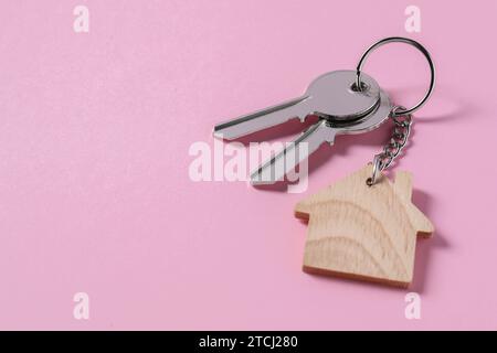 Metallische Schlüssel mit hölzernem Schlüsselanhänger in Form eines Hauses auf rosa Hintergrund, Platz für Text Stockfoto