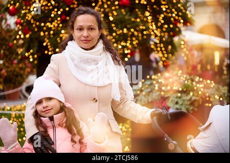 Schöne Mutter mit Kinderwagen und ihrer niedlichen Tochter, die in die Kamera schaut, draußen vor einem Weihnachtsbaum steht und festliche Atmosphäre genießt Stockfoto
