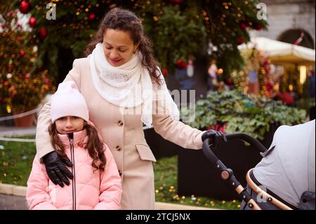 Mutter und Tochter gehen zusammen auf der Weihnachtsfeier. Glückliche Frau, die ihr Kind umarmt und den Kinderwagen schiebt, genießt die traditionelle Familienmarke Stockfoto