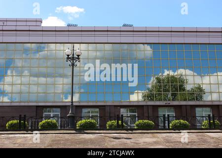 Grüner Baum und Wolken auf Einem blauen Himmel spiegeln sich in der Spiegelglaswand des Bürogebäudes mit grünen Büschen davor Stockfoto