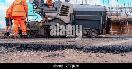 Asphaltfertiger mit heißem Asphalt, der neue Straßenoberflächen auf neuen Wohnbaustellen verlegt, und Straßenarbeiter in orangefarbenem Hi-viz als nächstes Stockfoto