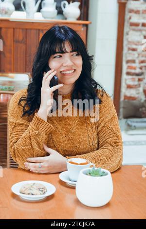 Vertikales Bild der jungen venezolanischen Latina mit schwarzen Haaren und braunem Pullover, sie sitzt lächelnd und telefoniert am Telefon, sitzt in Kaffee-Sho Stockfoto