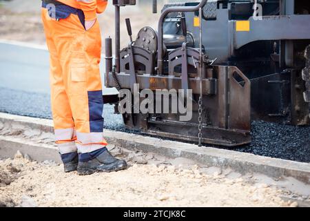 Asphaltfertiger mit heißem Asphalt, der neue Straßenoberflächen auf neuen Wohnbaustellen verlegt, und Straßenarbeiter in orangefarbenem Hi-viz als nächstes Stockfoto