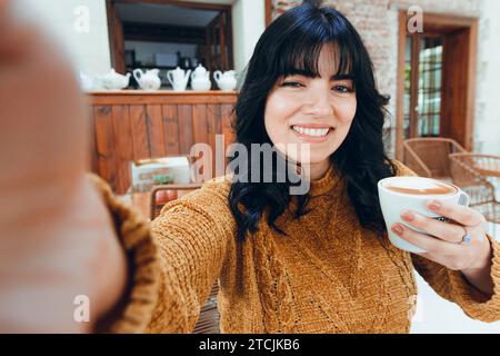 Selfie-Foto einer jungen lateinamerikanischen venezolanischen Frau, in braun, mit langen schwarzen Haaren, mit einer Tasse Kaffee in der Hand, glücklich sitzend und lächelnd und anschauend Stockfoto