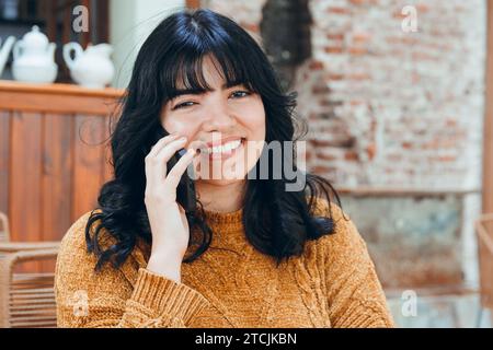 Vertikales Bild der jungen venezolanischen Latina mit schwarzen Haaren und braunem Pullover, sie sitzt lächelnd und telefoniert am Telefon, sitzt in Kaffee-Sho Stockfoto
