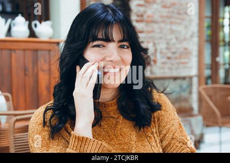 Vertikales Bild der jungen venezolanischen Latina mit schwarzen Haaren und braunem Pullover, sie sitzt lächelnd und telefoniert am Telefon, sitzt in Kaffee-Sho Stockfoto