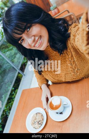Vertikales Bild einer jungen hispanischen Latina-Frau, die sehr glücklich lächelt und in die Kamera blickt, Selfie macht, sitzend im Café, Leute Konzept Kopierraum. Stockfoto