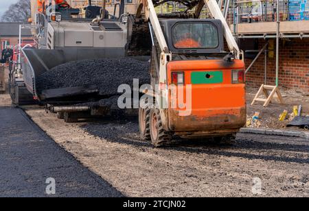 Asphaltfertiger mit heißem Asphalt, der neue Straßenoberflächen auf neuen Wohnbaustellen verlegt, und Straßenarbeiter in orangefarbenem Hi-viz als nächstes Stockfoto