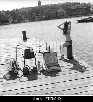 Lifestyle der 1950er Jahre. Eine junge Frau, die in der Sonne sitzt und einen Bikini trägt. Auf dem Bild befinden sich viele der zeitgemäßen Objekte aus Kunststoff, die zu dieser Zeit beliebt waren. Besonders bei Sommerferien oder Camping. Tassen und Untertassen auf dem Tisch. Ein praktischer Koffer für den Transport. 1958 Kristoffersson Ref. DB97-6 Stockfoto