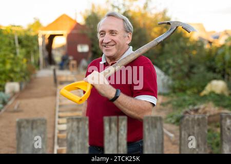 Ein erwachsener Mann stand entspannt neben dem Zaun, mit einer Schaufel im Garten, die lächelte Stockfoto