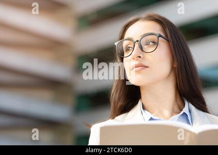 Die nachdenkliche europäische Millennial-Frau mit Brille schaut nach oben, hält Buch, Papier-Projekt offen Stockfoto