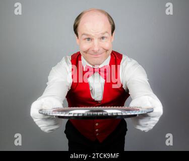 Porträt eines Kellners oder Dieners in weißen Handschuhen und roter Weste oder Weste, der freundlich lächelt und ein silbernes Serviertablett hält. Professionelle Gastfreundschaft. Stockfoto