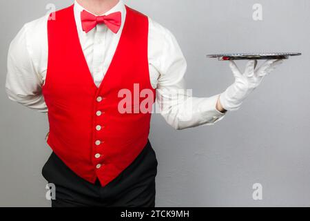 Porträt des Kellners in roter Weste oder Weste und roter Schleife mit silbernem Serviertablett. Konzept der Dienstleistungsbranche und des professionellen Gastgewerbes. Stockfoto