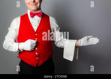 Porträt des Butlers oder Kellners in roter Weste und Schleife lächelnd mit Willkommensgeste. Konzept der Dienstleistungsbranche und professionelle Höflichkeit. Stockfoto