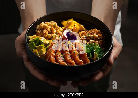 Die Hände eines Küchenchefs halten eine schwarze Schüssel, gefüllt mit einem farbenfrohen Arrangement einer Poke-Schüssel mit frischem Lachs, Reifen Avocadoscheiben und lebendigem Grün, g Stockfoto