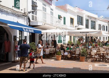 Touristen gehen durch eine enge, gemütliche Straße mit Restaurants und Geschäften im Dorf Fornells auf der spanischen Insel Menorca. Stockfoto