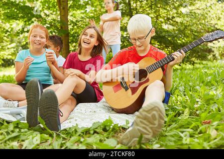 Glückliche Freundinnen, die neben dem Jungen sitzen und Gitarre spielen, während sie im Park auf einer Decke sitzen Stockfoto