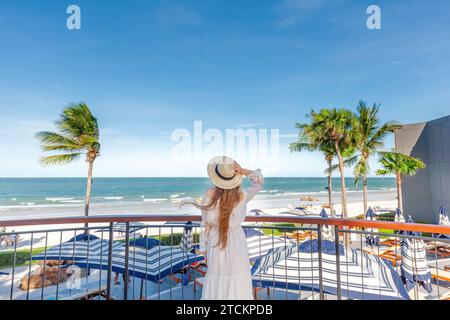 Blick auf die Küste mit einer Frau in weiß, die sich vom Balkon des Resorts an einem Strohhut festhält Stockfoto