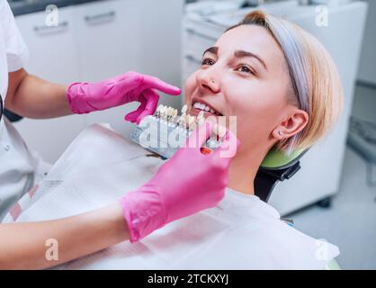 Junge Frau, die im Stomatologie-Klinikstuhl sitzt und lächelnd ihre Zähne zeigt. Zahnarzt, der Zahnfarben mit Farbtabellen wählt. Gesundheitswesen, Mout Stockfoto