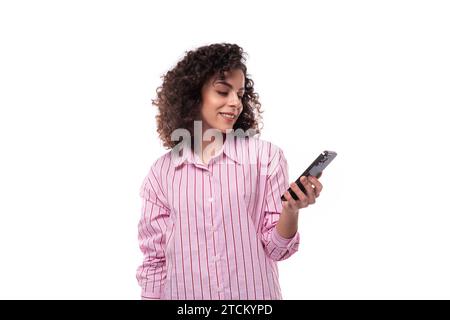 Eine junge selbstbewusste Brünette lockige Geschäftsfrau in einem gestreiften rosa Hemd schreibt eine Nachricht am Telefon Stockfoto