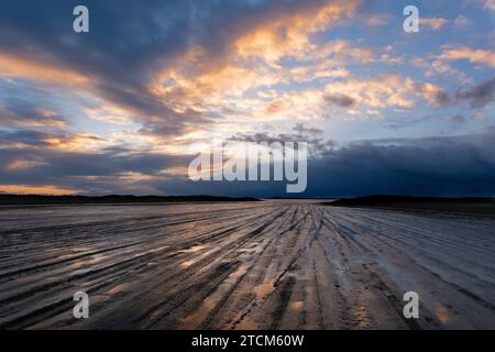 Spuren von Autoreifen an einem matschigen Strand, der bei Einbruch der Dunkelheit zum Horizont führt, unter einem wunderschön farbigen Himmel, bedrohliche Wolken in der Ferne Stockfoto
