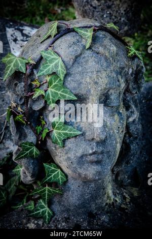 Eine alte Sandsteinstatue auf einem mit Efeu bedeckten Friedhof. Ein Efeuhaarkranz auf dem Kopf einer Sandsteinskulptur. Winter. Friedhof. Stockfoto