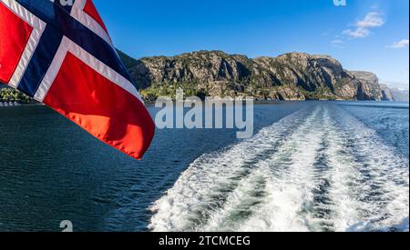 Bandera noruega en un barco ondeando al viento y el paisaje del Fiordo de la Luz o Lysefjord con la estela del barco, Stavanger, Noruega Stockfoto