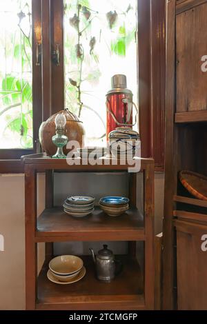 Innenraum der vietnamesischen Küche mit Porzellanvase, Aluminiumkessel, blauer Porzellanplatte, Rattanuntersetzer, Öllampe, Vintage Thermosflasche in Stockfoto